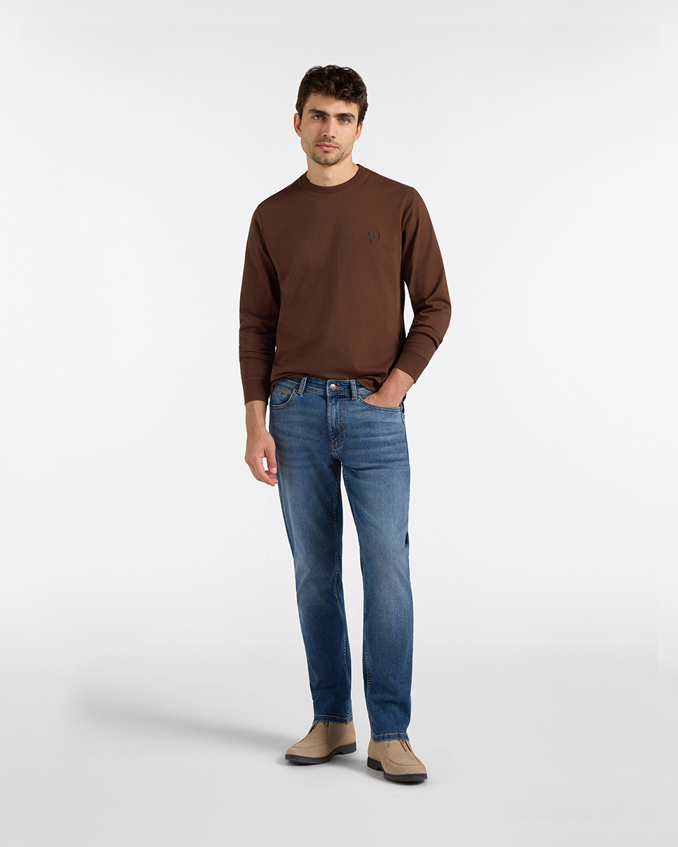 Per la categoria pantaloni HB Jeans, un modello indossa un paio di jeans blu lavaggio medio a gamba dritta, abbinati a una maglia a maniche lunghe marrone scuro e scarponcini scamosciati beige su sfondo bianco.
