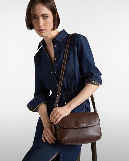 Una donna con capelli corti castani indossa un abito in denim blu scuro a maniche lunghe e porta a tracolla una borsa in pelle marrone scuro con patta e risvolto.