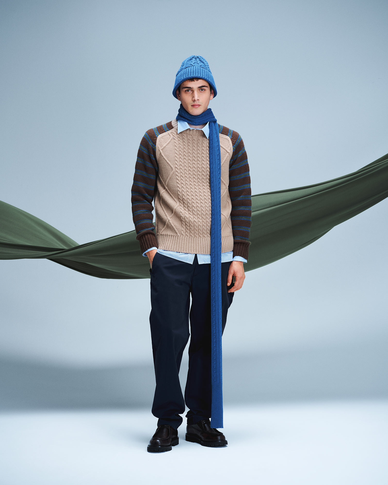Uomo con maglione intrecciato beige, pantaloni scuri e berretto azzurro con sciarpa coordinata