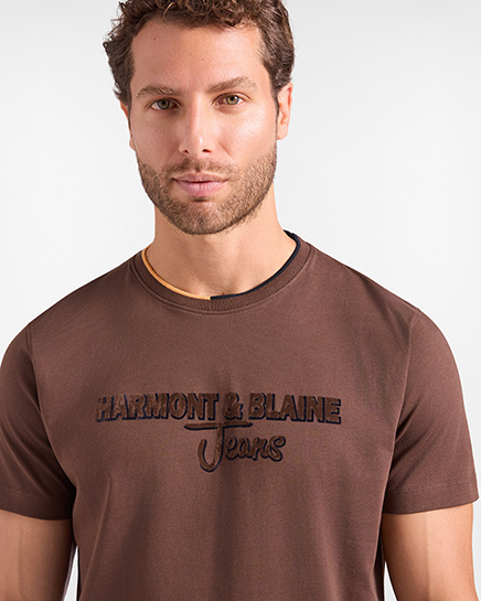 Per la categoria t-shirts HB Jeans, un modello indossa una t-shirt a maniche corte di colore marrone scuro con girocollo caratterizzato da un dettaglio di profilo a contrasto e il logo "HARMONT & BLAINE Jeans" ricamato sul petto.