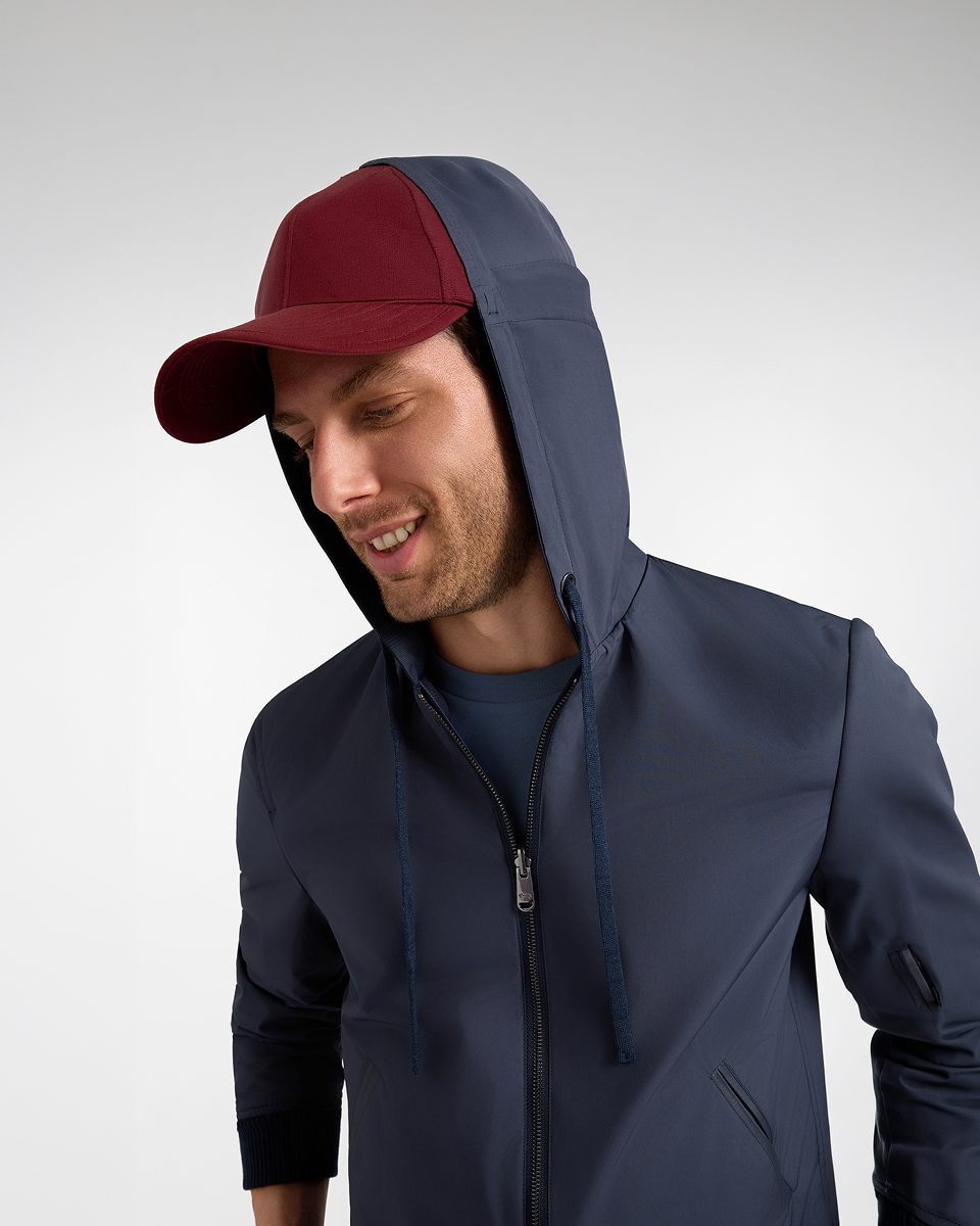 Uomo sorridente che indossa una giacca impermeabile blu scuro con cappuccio e un cappellino con visiera di colore bordeaux sotto il cappuccio.
