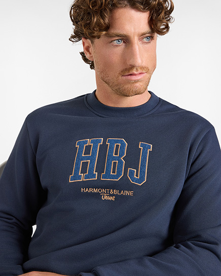 Un uomo con capelli ricci castani e occhi azzurri indossa una felpa girocollo di colore blu scuro, con un grande logo ricamato sul petto. Le lettere "HBJ" sono in stile college, di colore blu con contorno arancione, e sotto &egrave; presente il testo "HARMONT&BLAINE Jeans" ricamato in arancione. Questo capo appartiene alla categoria felpe HB Jeans.