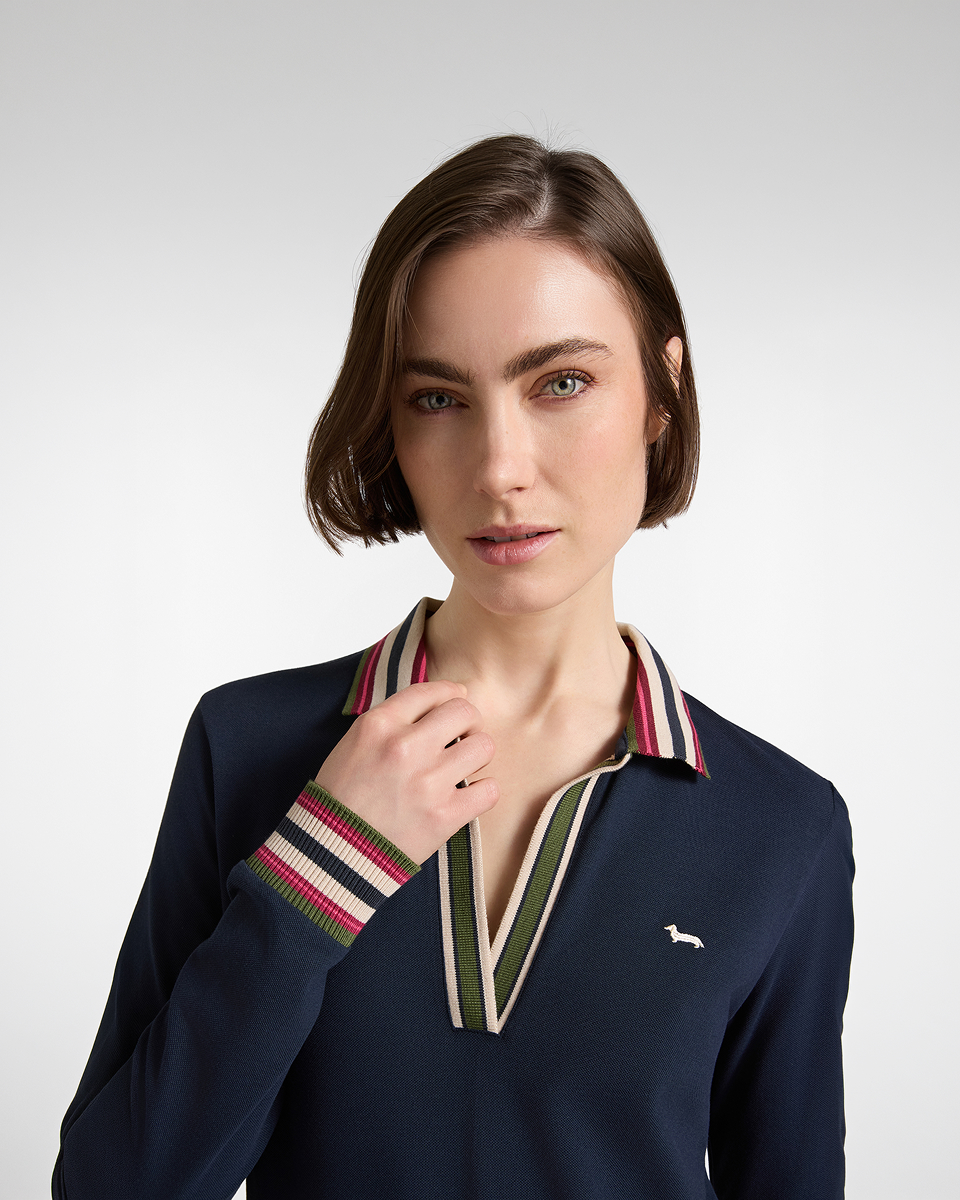 Un primo piano di una donna con capelli corti castani che indossa una polo a maniche lunghe blu scuro con scollo a V, colletto e polsini rifiniti a righe multicolore e piccolo logo ricamato sul petto.