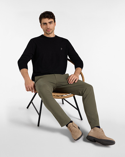 Un uomo siede su una sedia in posa rilassata mentre indossa un maglione a maniche lunghe nero e un paio di pantaloni chino di colore verde oliva. Il look &egrave; completato da un paio di scarponcini scamosciati di colore beige con suola nera. Lo sfondo &egrave; bianco. Questo outfit fa parte della categoria pantaloni HB Jeans.