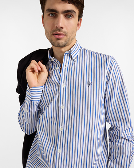 Per la categoria camicie HB Jeans, un modello indossa una camicia a righe verticali bianche, blu e grigie con colletto button-down e logo ricamato sul petto, tenendo una giacca nera sulla spalla su sfondo bianco.