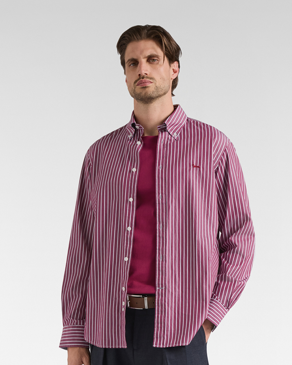 Un uomo castano con la barba corta indossa una camicia a maniche lunghe sbottonata a righe verticali bianche e bordeaux, sopra una maglietta bordeaux e pantalone scuro con cintura.