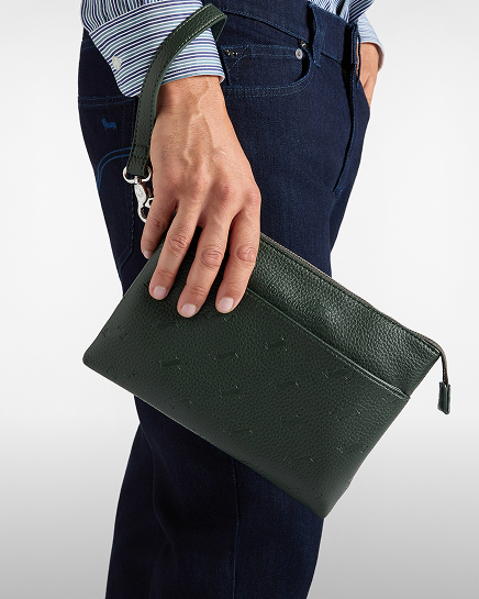 Un primo piano di una persona con camicia a righe e jeans scuri che tiene in mano una pochette in pelle verde scuro con laccetto da polso e chiusura a zip.