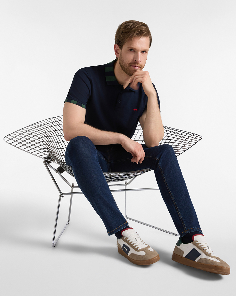 Un uomo biondo con la barba, seduto su una sedia con struttura in rete d'acciaio cromato, indossa una polo blu scuro a maniche corte con colletto a righe verdi e blu scuro e polsini a righe, jeans blu scuro e scarpe da ginnastica grigio scuro e beige.