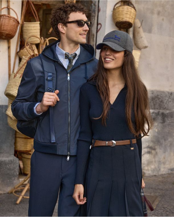 L'immagine ritrae una coppia che cammina sorridente all'esterno di una bottega artigianale decorata con cesti in vimini appesi alle pareti. L'uomo indossa occhiali da sole scuri, una giacca a vento blu scuro con cappuccio sopra una camicia a righe e cravatta, un pantalone abbinato e uno zaino in spalla. La donna, al suo fianco, indossa una maglia a coste blu scuro a maniche lunghe con scollo a V, una gonna a pieghe coordinata con cintura in pelle marrone e un cappello da baseball grigio con logo ricamato blu.