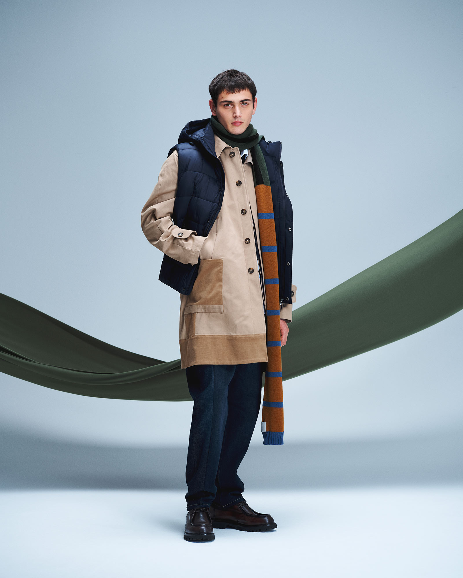 Uomo con parka beige e gilet imbottito blu, sciarpa a righe e pantaloni scuri