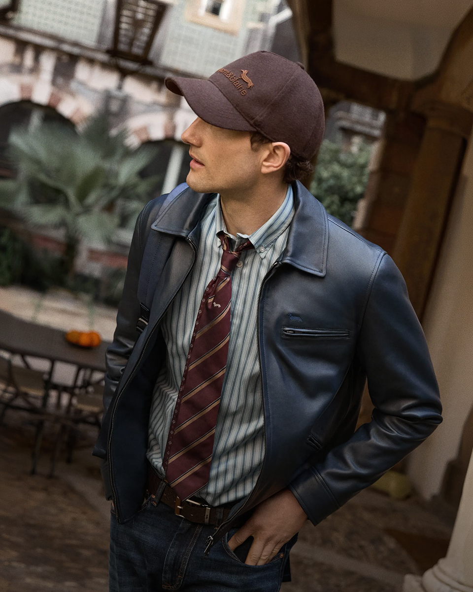 Un uomo visto di profilo indossa un giubbotto in pelle blu scuro, una camicia a righe verticali con cravatta bordeaux a righe, cappello da baseball marrone con logo e jeans.