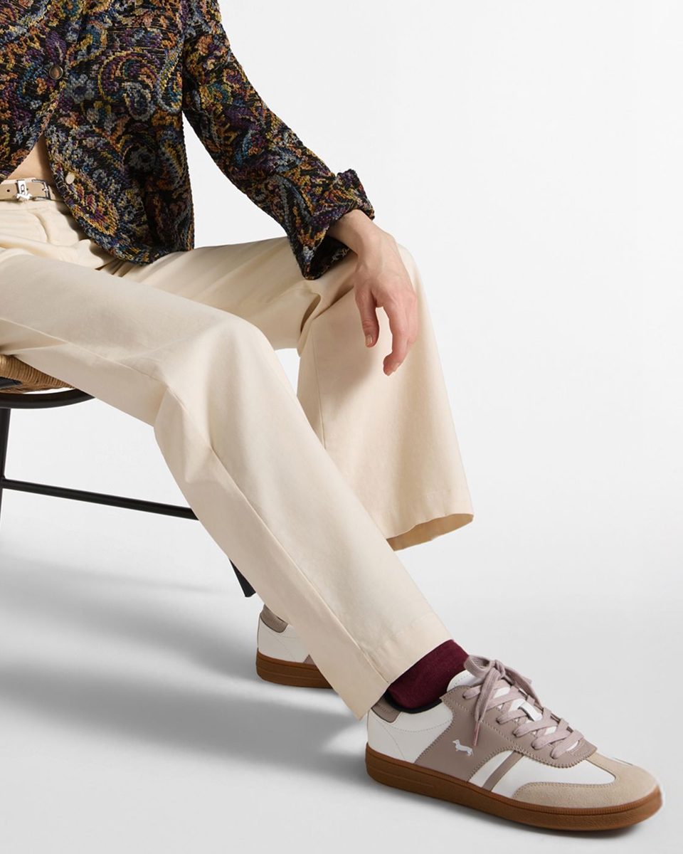 Una persona seduta su una sedia in vimini indossa pantaloni ampi color panna e sneaker da donna bianche con dettagli beige, rosa cipria e suola in gomma color caramello.